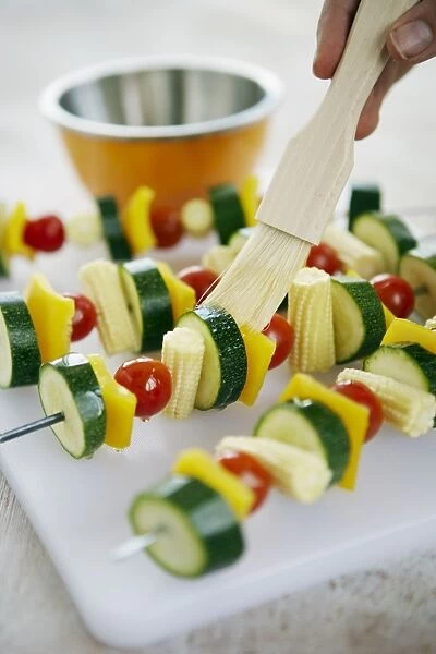 Using basting brush to apply sunflower oil to raw vegetable kebabs on skewers on plastic chopping board in kitchen