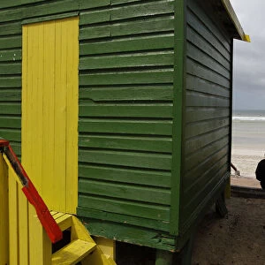 A spectator sits between beach huts as surfers prepare to take to the water in an