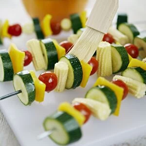 Using basting brush to apply sunflower oil to raw vegetable kebabs on skewers on plastic chopping board in kitchen