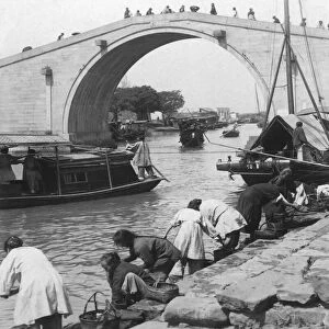 Historical image in black and white of Chinese people working along a river with Junk boats and bridge, circa 1900; China